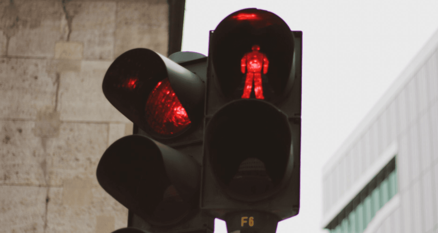 A close-up of a pedestrian traffic light displaying a glowing red standing figure, with a standard red traffic light illuminated beside it. Set against a blurred urban building background, the image serves as a metaphor for cognitive inhibition, self-control, and the ability to pause impulsive actions.